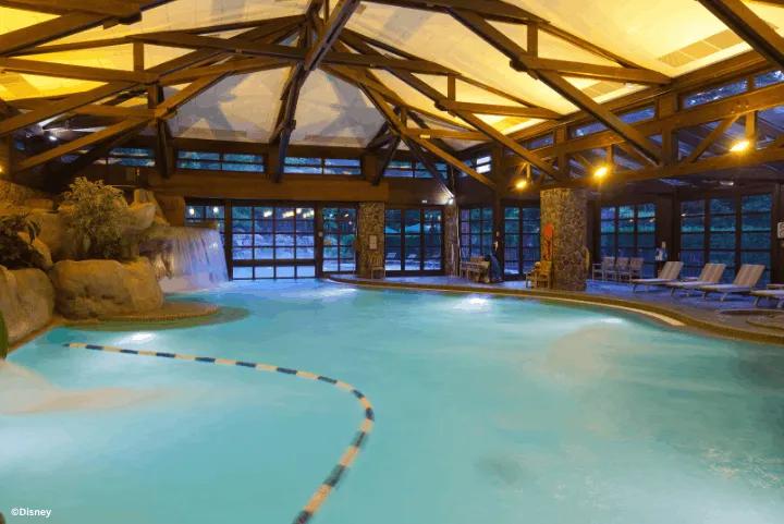 An indoor swimming pool featuring exposed wooden beams, a stone waterfall, and lounge chairs
