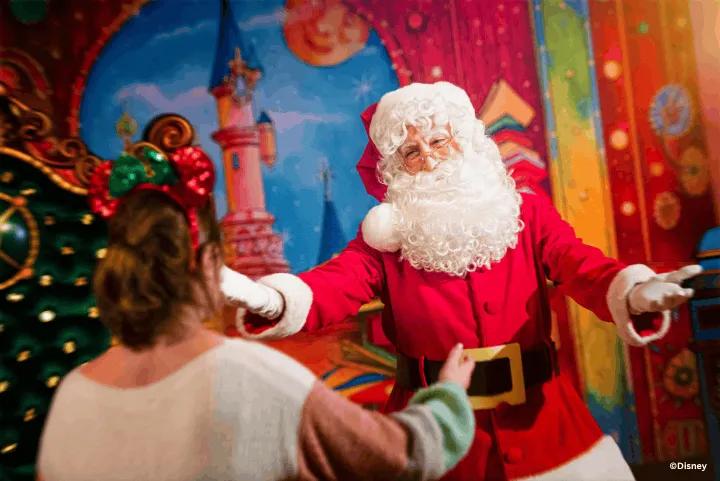 A guest wearing festive Minnie Mouse ears walks toward Santa Claus, who is standing with open arms in front of a brightly painted, magical backdrop