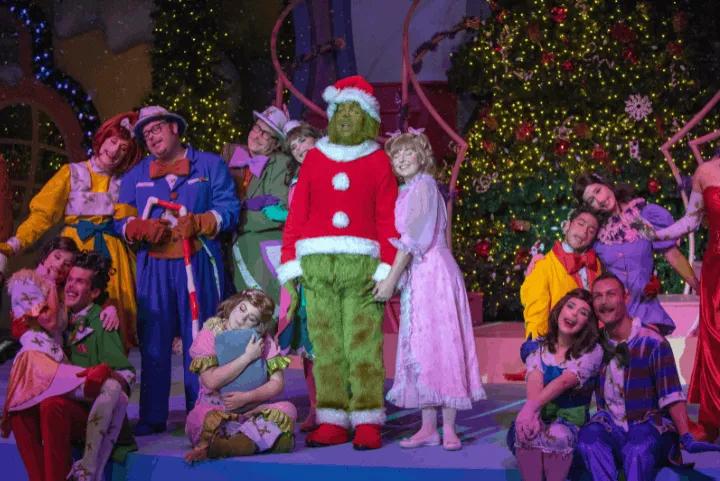 The Grinch stands on stage in a Santa suit alongside Cindy Lou Who and other festively dressed characters