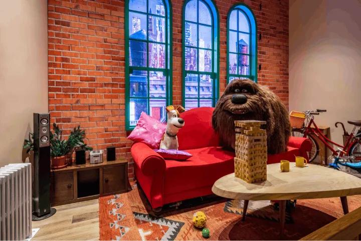 Two animatronic dog characters, Max and Duke, sitting on a red couch near a Jenga tower in an apartment setting