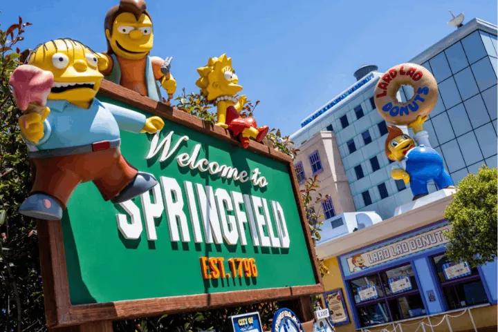 A Welcome to Springfield sign featuring Simpsons character statues, with the Lard Lad Donuts shop in the background