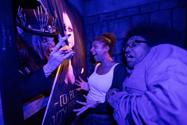 Two frightened guests reacting to a monstrous scareactor wearing a skull-adorned top hat inside the blue-lit Ozzy Osbourne haunted house, standing next to a poster reading "God Bless Ozzy Osbourne"