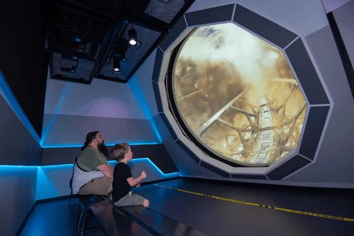 A man and child sitting in the Spaceport KSC Observation Bay watching a simulated rocket launch