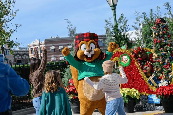 The Earl the Squirrel mascot, wearing a green sweater and a plaid hat, interacts with guests next to a decorated Christmas tree and sleigh