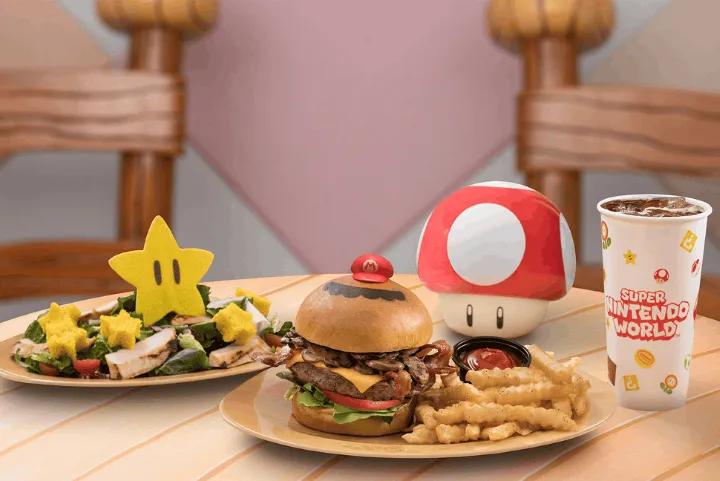 A table featuring themed food items from Toadstool Cafe, including a star-themed salad, a burger topped with a Mario hat pick, a mushroom-shaped soup bowl, and a branded beverage cup