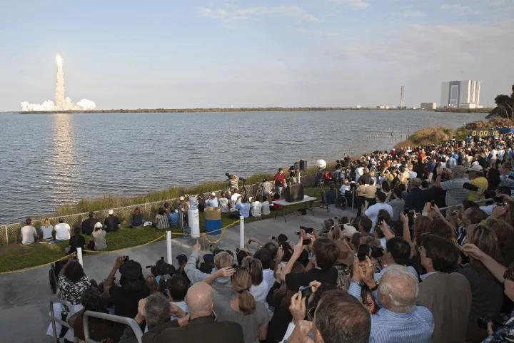 A large crowd gathered at a park watching a rocket launch across the water