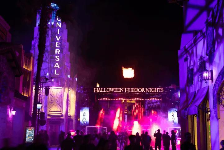 A crowd of guests walking toward the brightly lit Halloween Horror Nights entrance gate with bursts of fire in the night sky