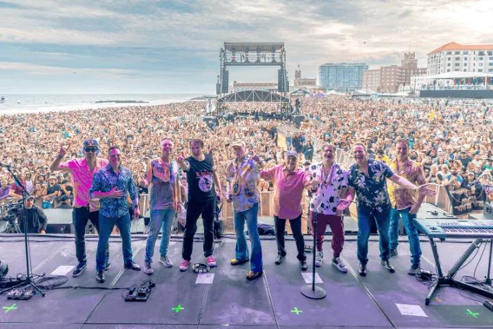 The Beach Boys standing together on stage in front of a large festival crowd on a beach