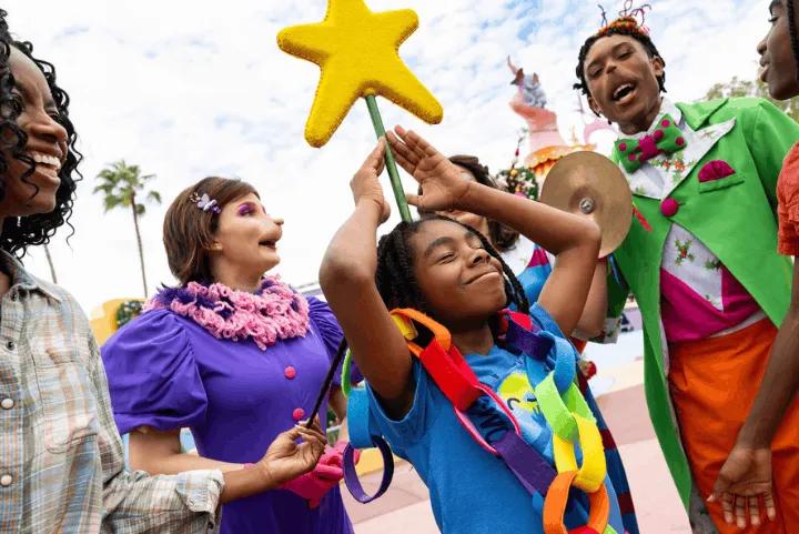 A young child wearing a colorful paper chain smiles with their eyes closed while holding a yellow star on a stick, surrounded by costumed Whoville characters