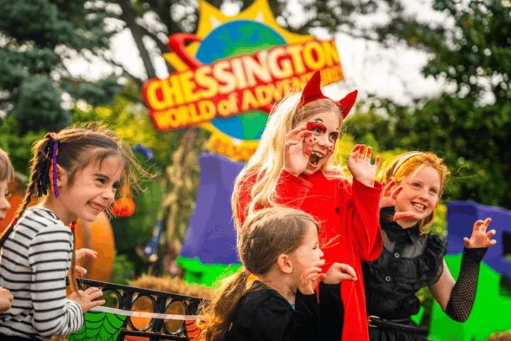 Three young girls dressed in Halloween costumes smiling and posing playfully in front of the Chessington World of Adventures entrance sign