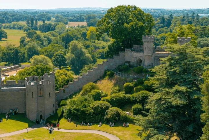 An elevated view of the stone walls and towers of Warwick Castle surrounded by lush green trees and lawns, with visitors scattered across the grass below