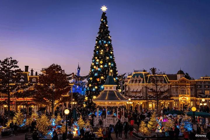 A tall, illuminated Christmas tree stands in a crowded town square at dusk, surrounded by festive buildings and a decorated gazebo