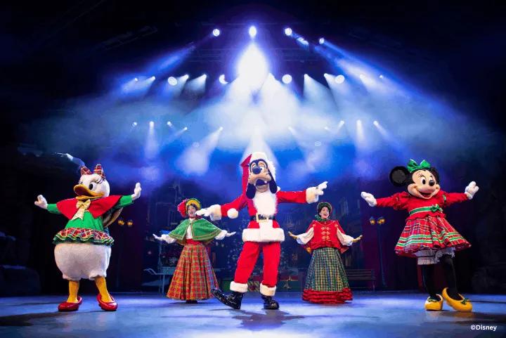 Daisy Duck, Goofy dressed as Santa Claus, and Minnie Mouse performing a holiday show on a brightly lit stage alongside two festive carolers