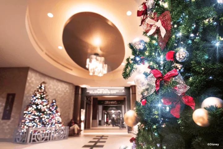 A Christmas tree decorated with red bows and metallic ornaments in a modern hotel lobby, with another glowing tree visible in the background