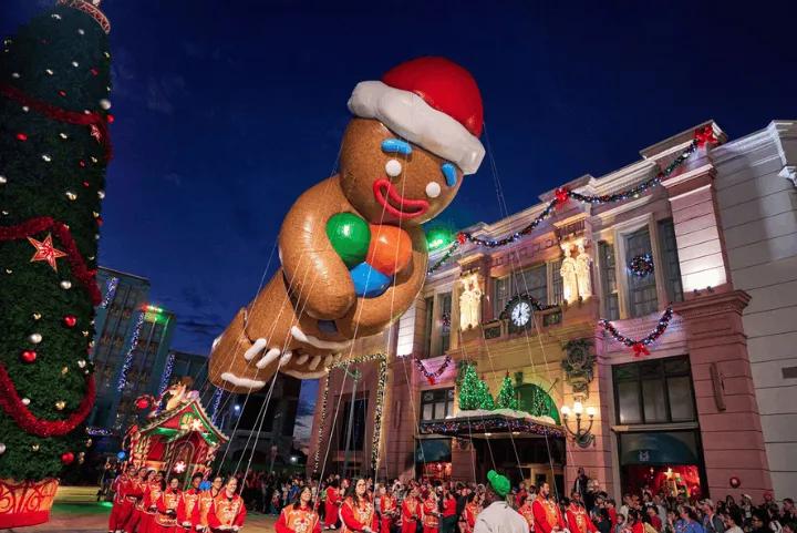 A giant Gingerbread Man balloon wearing a Santa hat floats above a street at night during a holiday parade