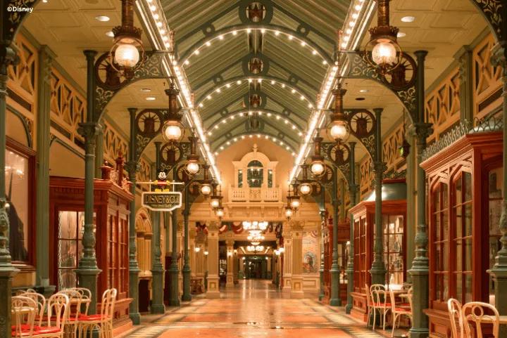 An ornate indoor walkway with lit arched ceilings, hanging lanterns, and vintage shop facades