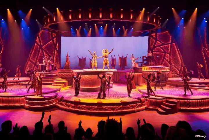 Performers in vibrant costumes acting and dancing on a lit, tiered stage in front of an audience