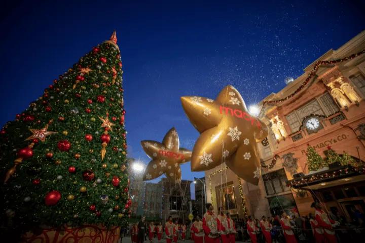 Two giant gold star balloons featuring the Macy's logo float past a tall decorated Christmas tree and city street facades during a nighttime holiday parade