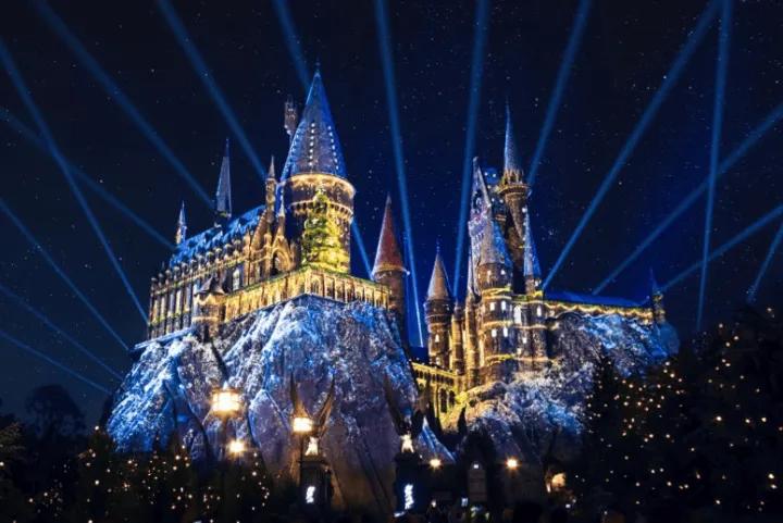 Hogwarts Castle illuminated with holiday light projections and searchlights against a dark night sky