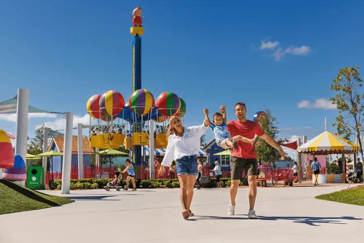 A mother and father swinging their young son by his arms while walking in front of a Peppa Pig balloon ride at a theme park