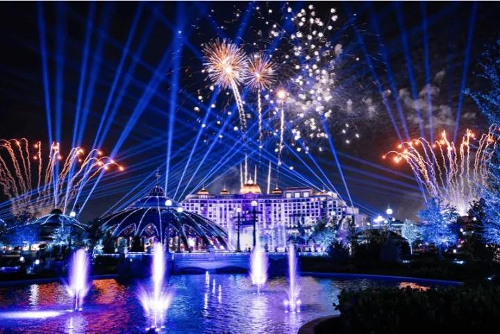 A nighttime theme park spectacular featuring bright fireworks, blue spotlights, and illuminated fountains in front of a grand hotel