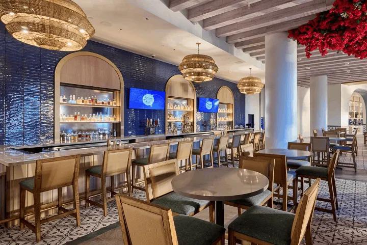 A vibrant bar and lounge area featuring a blue textured wall, wooden stools, overhead woven light fixtures, and red floral ceiling decor