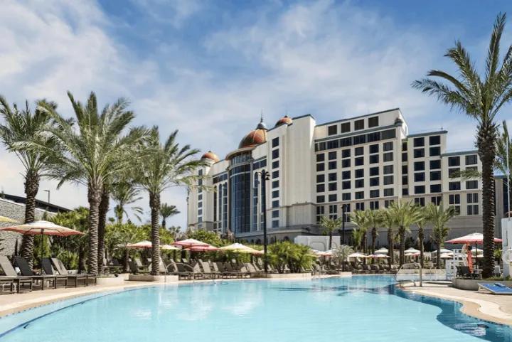 Outdoor view of the Universal Helios Grand Hotel exterior and large resort pool area with palm trees, lounge chairs, and umbrellas under a daytime sky
