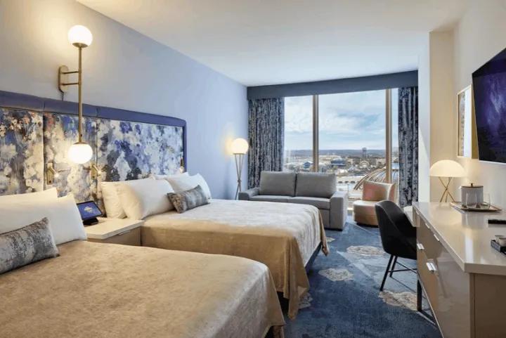 A standard hotel room featuring two queen beds with blue and purple floral patterned headboards, a sofa, and large windows with outdoor views