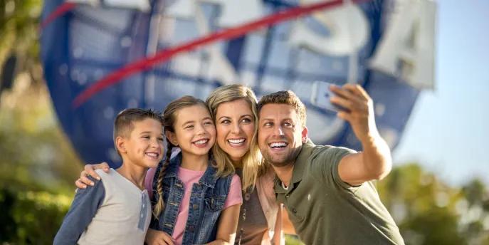 Kennedy Space Center Family of four, a man holding up a phone to take a selfie with wife and two kids, one boy and one girl. They are stood in front of a large sign, shaped like the earth, circular with red stripes over and the letters in white spelling NASA.