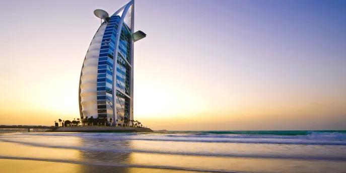 Burj Al Arab Sail shaped building coloured white with many windows and a helicopter pad on top. It is in the middle of the ocean on a man made island, with one road connecting it with mainland.