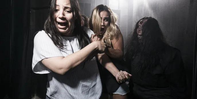 Howl-O-Scream Two young women in a dark room with a bright light shining behind with terrified facial expressions, clutching onto one another and dodging another person who is lurking in the dark. The third person has black hair and a wound on their head which seems to be bleeding down their face. Their hair is messy and they are pulling a scary face with their tongue poking out.