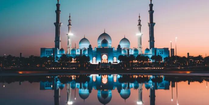 A beautiful religious temple in Abu Dhabi located overlooking a lake. The sun is setting with a pink and blue hue in the sky and the temple is perfectly reflected in the lake. It is a white building with three white domes and four big pillars sticking out.