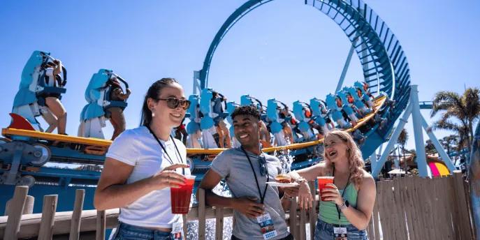 Three young adults stood in front of the Pipeline rollercoaster drinking cocktails and eating festival plates