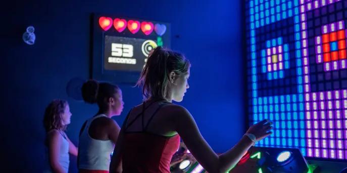 Activate Orlando Girl pressing a gaming pad in front of a screen with a game on it. There are two other players and on the wall it displays their lives left.