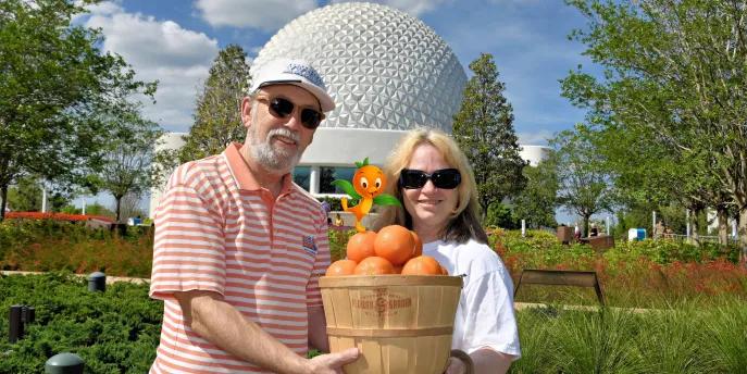 Man and woman holding a basket full of oranges in front of the giant golf ball looking structure and green grass and trees.