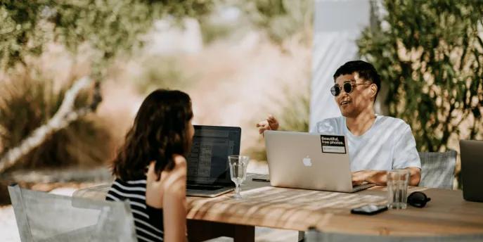 Workation A woman and man sat at a wooden table outside talking in the sun with their work laptops open.