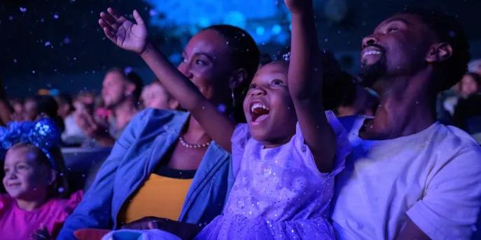 Frozen Sing Along Celebration A little girl smiling and holding her arms up while sat with her mum and dad in a theatre