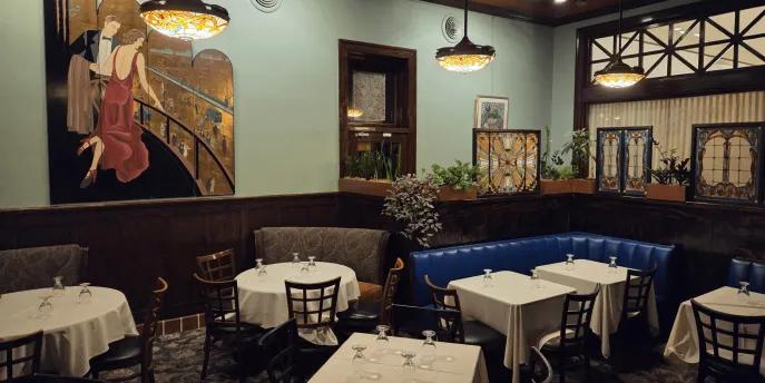 The Chef’s Table at The Edgewater Large blue room with dark decor, paintings and tables with white tablecloths inside.
