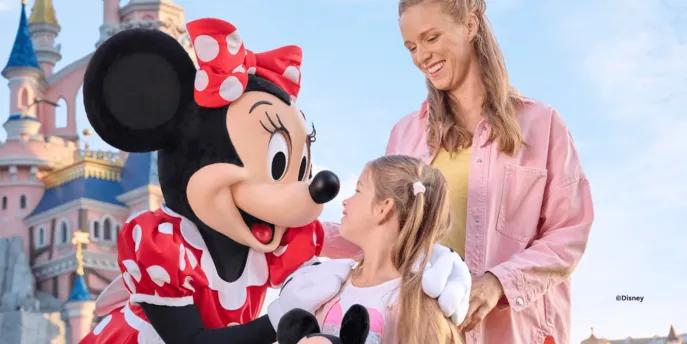 A young girl and her mother smiling with the Minnie Mouse character in front of Sleeping Beauty Castle at Disneyland Paris.