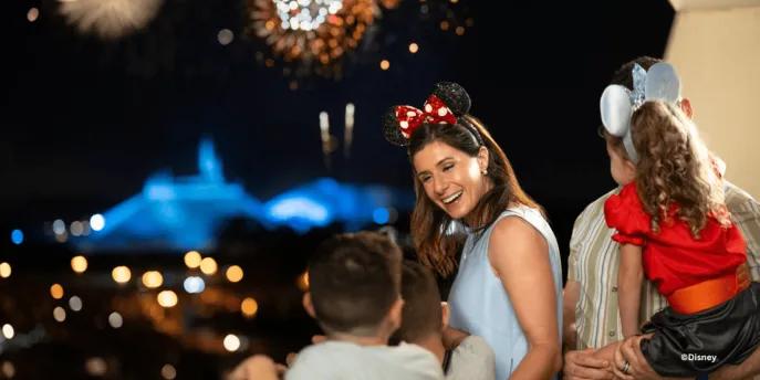 A family wearing Mickey and Minnie ears watches a spectacular nighttime fireworks display over a lit-up theme park at Walt Disney World Resort.