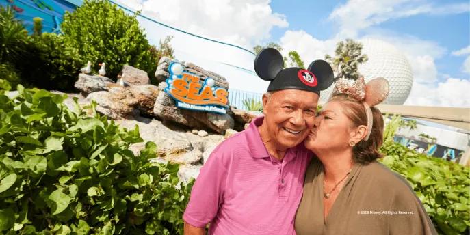 Couple wearing Mickey and Minnie ears near The Seas with Nemo & Friends pavilion at EPCOT, with Spaceship Earth in the background