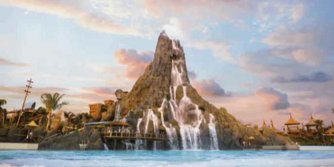 A wide shot of a massive artificial volcano with cascading waterfalls flowing into a large, wavy blue pool, under a pastel pink and blue sunset sky with scattered clouds