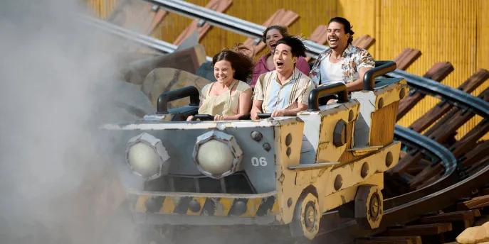 Riders smiling and cheering aboard a rustic mine cart roller coaster emerging from a cloud of mist