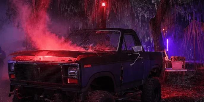 A dark pickup truck with red smoke rising from the open hood parked in swampy woods filled with neon lighting