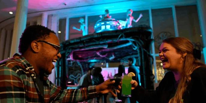Two guests laughing and clinking colorful themed cocktails at a Universal Orlando Resort hotel Halloween Horror Nights bar experience
