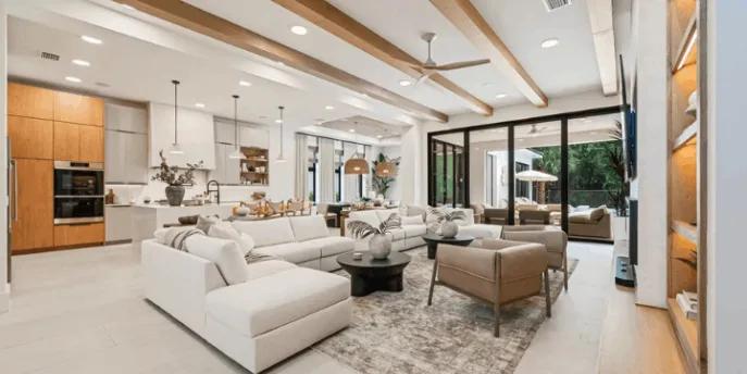 A spacious modern open-concept living room and kitchen featuring wooden ceiling beams, a large white sectional sofa, and floor-to-ceiling glass patio doors