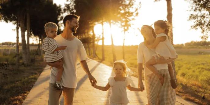 A happy family of five holding hands and carrying young children while walking along a paved path at sunset