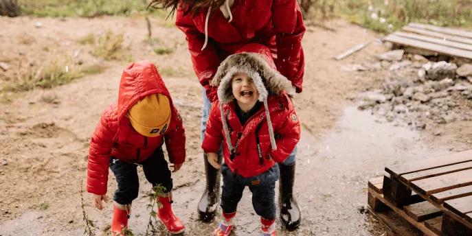 Two young children in red raincoats laugh and play in a muddy puddle while an adult in a red coat bends down beside them, illustrating a rainy day out with the family