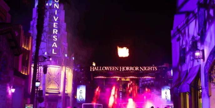 A crowd of guests walking toward the brightly lit Halloween Horror Nights entrance gate with bursts of fire in the night sky