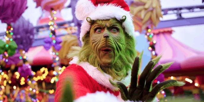 The Grinch, wearing a red Santa suit and hat, poses with a surprised expression in a brightly decorated theme park area